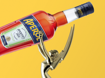 Emmy award holding bottle of oversized Aperol on. ayellow background